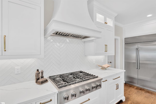 kitchen with decorative backsplash, custom exhaust hood, glass insert cabinets, light stone counters, and appliances with stainless steel finishes