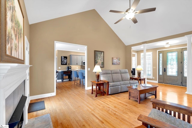 living room featuring light wood finished floors, high vaulted ceiling, a ceiling fan, decorative columns, and a fireplace with flush hearth