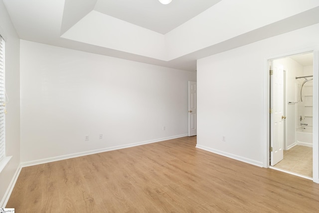 unfurnished bedroom with light wood-style floors, a raised ceiling, baseboards, and ensuite bath