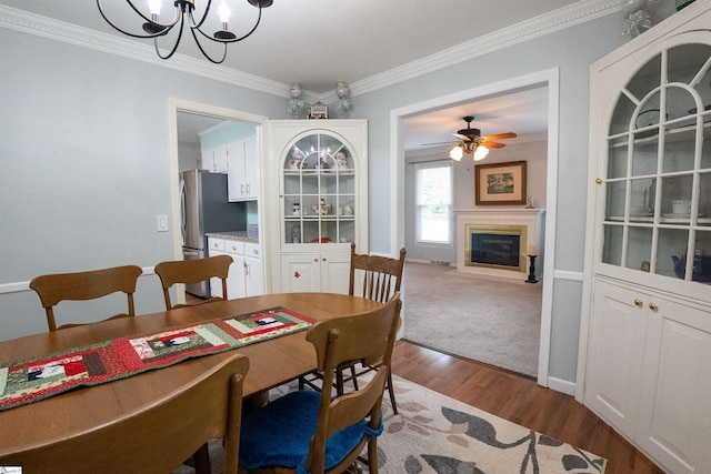 dining area with wood finished floors, baseboards, ceiling fan with notable chandelier, a glass covered fireplace, and ornamental molding