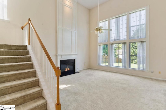 unfurnished living room with a fireplace, a high ceiling, stairway, a ceiling fan, and carpet
