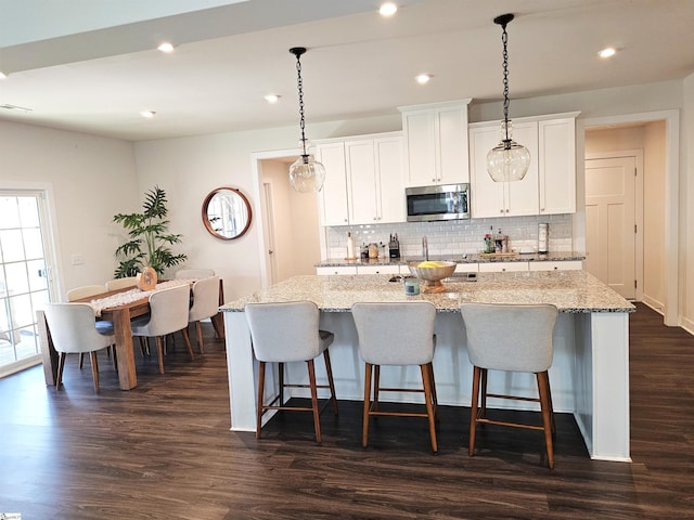 kitchen featuring decorative backsplash, a kitchen island with sink, dark wood-style flooring, white cabinets, and stainless steel microwave