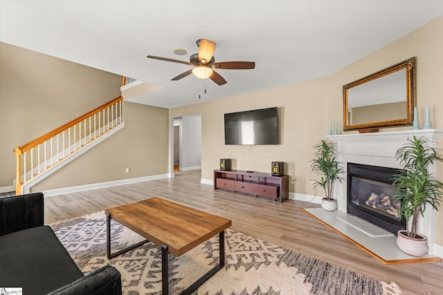 living room featuring a tiled fireplace, a ceiling fan, baseboards, wood finished floors, and stairway