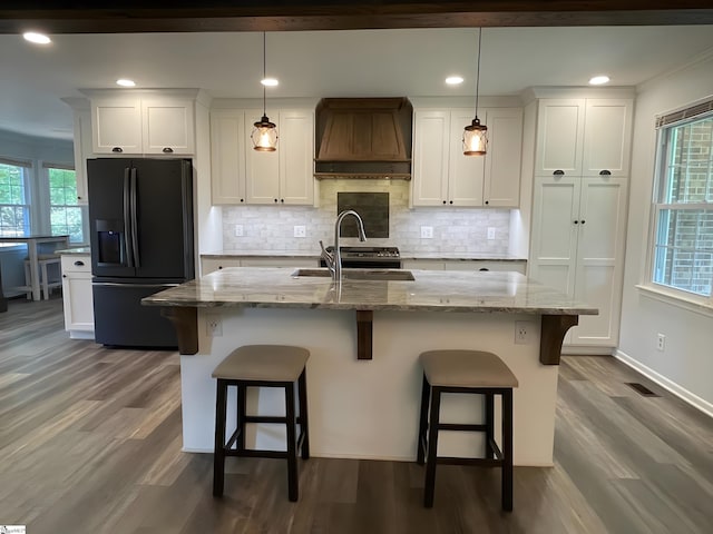 kitchen with black fridge, a sink, premium range hood, white cabinetry, and backsplash