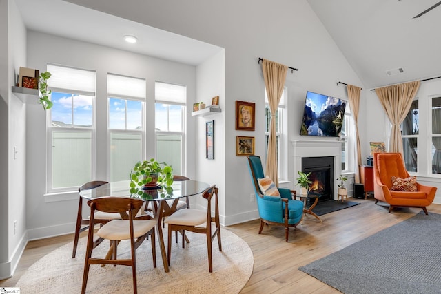 dining room featuring visible vents, high vaulted ceiling, baseboards, a warm lit fireplace, and light wood-style flooring