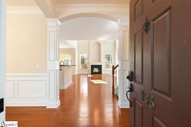 foyer entrance with crown molding, wood-type flooring, arched walkways, a glass covered fireplace, and ornate columns