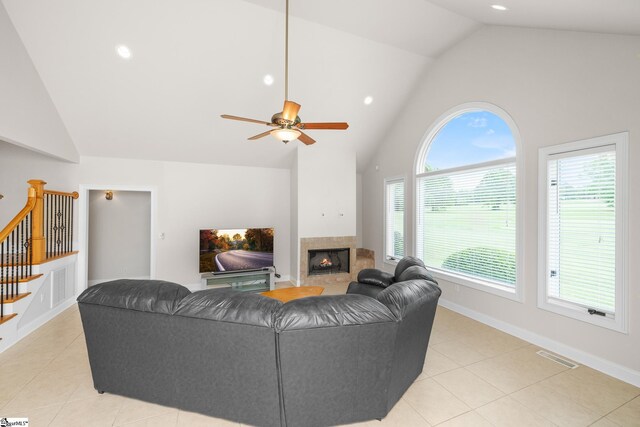 living room featuring stairs, light tile patterned flooring, ceiling fan, a warm lit fireplace, and high vaulted ceiling
