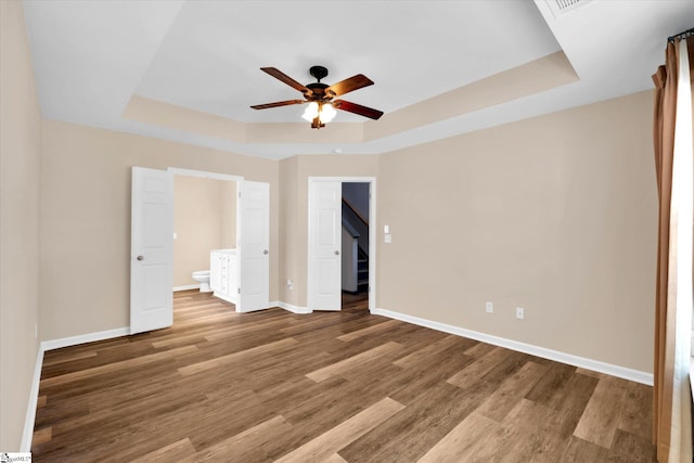 unfurnished bedroom featuring wood finished floors, baseboards, a tray ceiling, a ceiling fan, and ensuite bath
