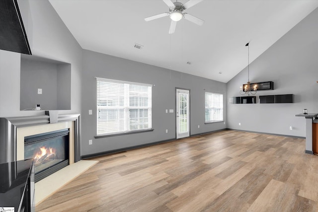 unfurnished living room with visible vents, high vaulted ceiling, ceiling fan with notable chandelier, light wood finished floors, and a glass covered fireplace