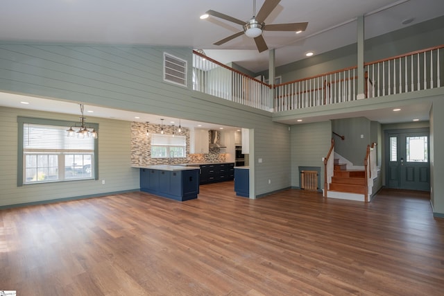 unfurnished living room with stairs, ceiling fan with notable chandelier, wood finished floors, and high vaulted ceiling