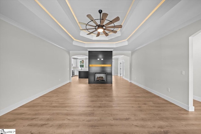 unfurnished living room featuring ceiling fan, crown molding, arched walkways, light wood-type flooring, and a tray ceiling