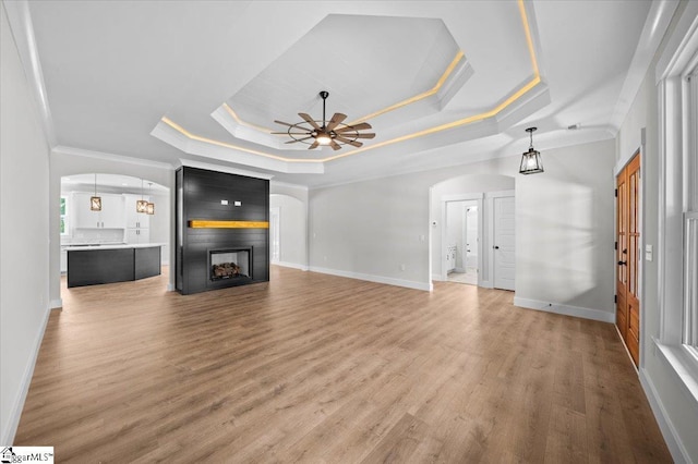 unfurnished living room featuring ceiling fan, arched walkways, a raised ceiling, light wood finished floors, and ornamental molding