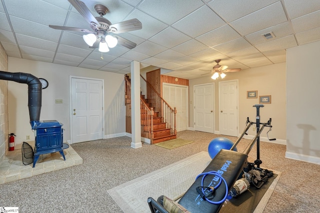 exercise room with a paneled ceiling, a wood stove, ceiling fan, and carpet floors