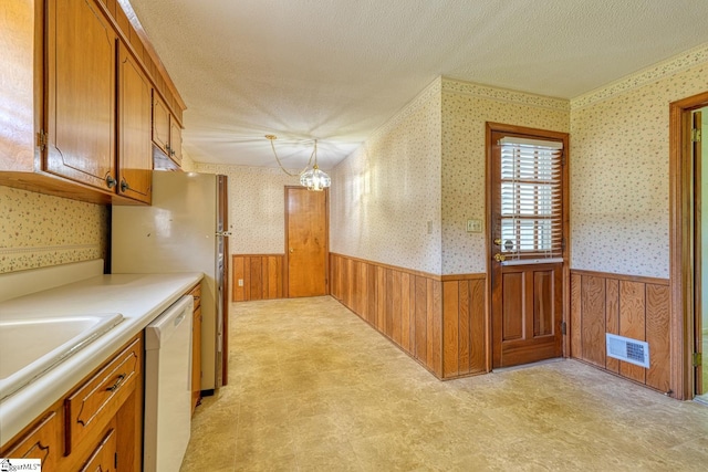 kitchen with brown cabinets, a textured ceiling, dishwasher, wainscoting, and wallpapered walls