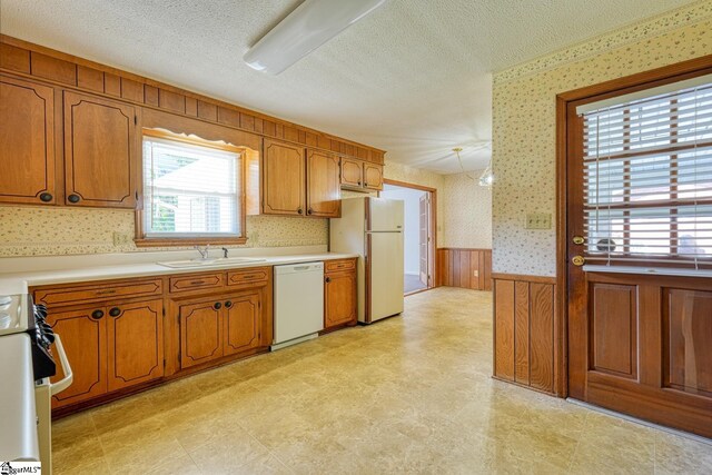 kitchen with a sink, white appliances, light floors, wainscoting, and wallpapered walls