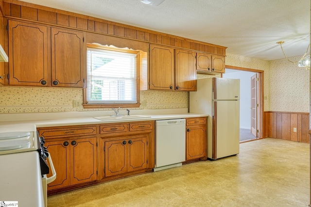 kitchen with wallpapered walls, brown cabinetry, white appliances, wainscoting, and a sink