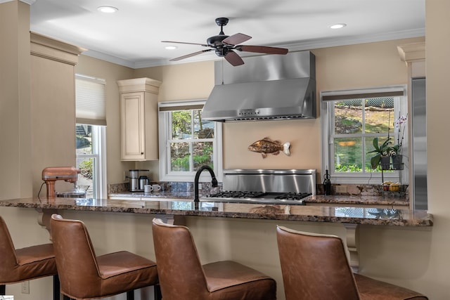 kitchen featuring wall chimney range hood, dark stone counters, a breakfast bar, ceiling fan, and ornamental molding