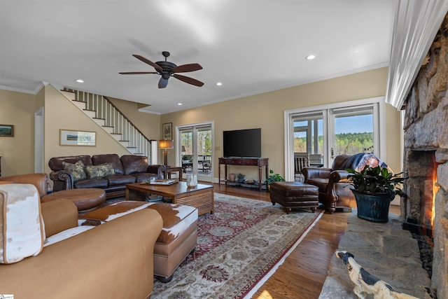 living room featuring a stone fireplace, healthy amount of natural light, wood finished floors, stairs, and ornamental molding