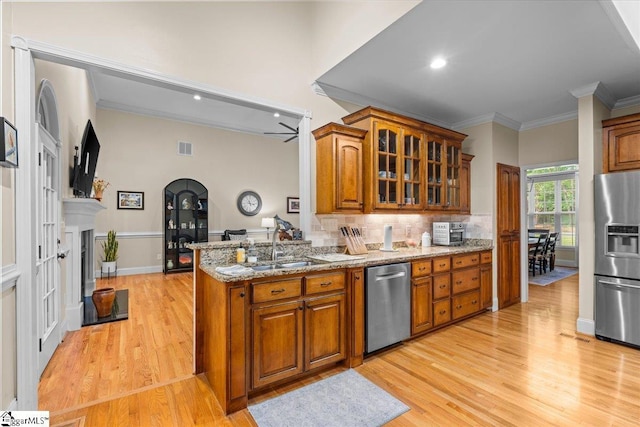 kitchen with appliances with stainless steel finishes, brown cabinetry, light wood finished floors, light stone counters, and glass insert cabinets