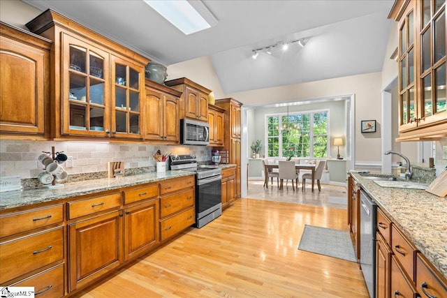 kitchen featuring appliances with stainless steel finishes, a sink, vaulted ceiling, brown cabinetry, and light wood-type flooring