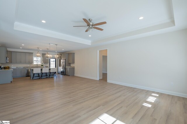 unfurnished living room featuring a raised ceiling, ceiling fan, light wood finished floors, recessed lighting, and ornamental molding