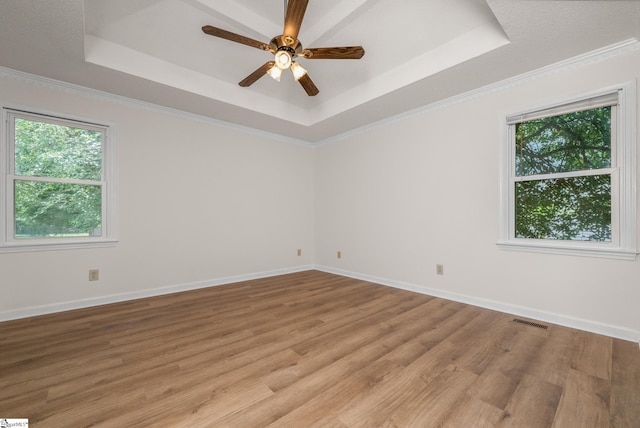 empty room with a tray ceiling, healthy amount of natural light, light wood-style flooring, a ceiling fan, and crown molding