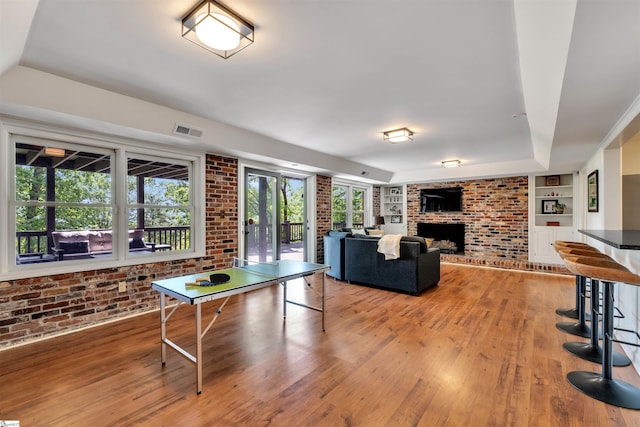 recreation room featuring brick wall, built in shelves, light wood-type flooring, a fireplace, and a tray ceiling