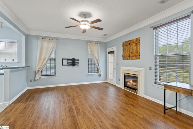 unfurnished living room with a ceiling fan, wood finished floors, healthy amount of natural light, a glass covered fireplace, and crown molding