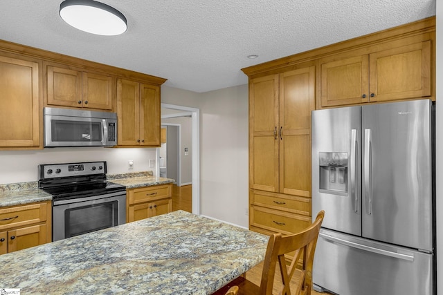 kitchen with appliances with stainless steel finishes, a textured ceiling, brown cabinetry, and light stone countertops