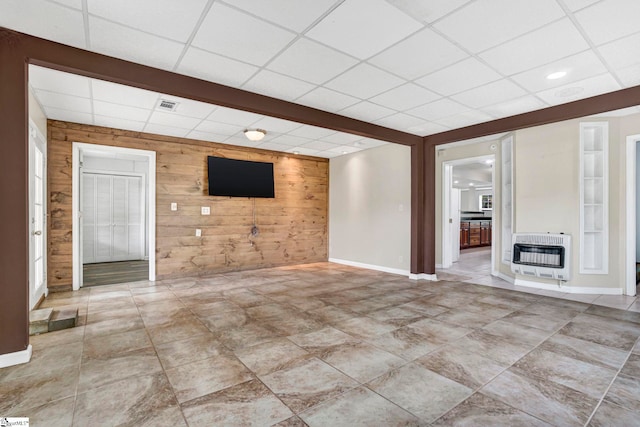 unfurnished living room with heating unit, a drop ceiling, wooden walls, and baseboards