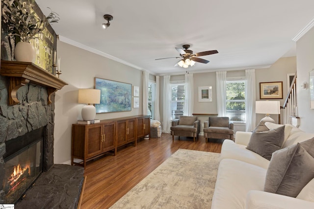 living room featuring a ceiling fan, wood finished floors, ornamental molding, a stone fireplace, and stairway