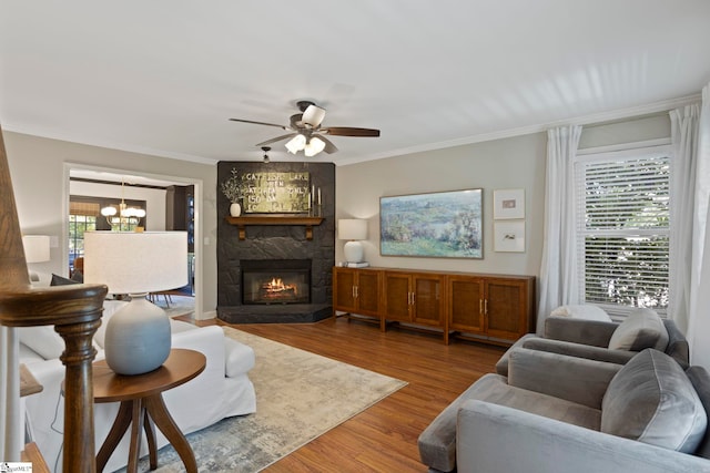 living area with a chandelier, a ceiling fan, crown molding, a stone fireplace, and wood finished floors