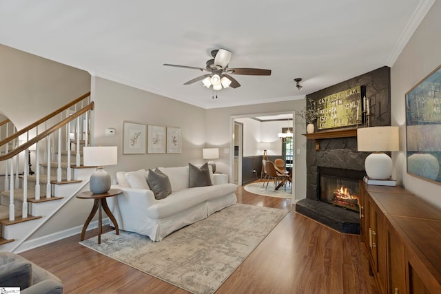 living room featuring stairs, a ceiling fan, wood finished floors, crown molding, and a stone fireplace