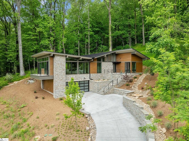 mid-century home with driveway, stone siding, an attached garage, and stairs