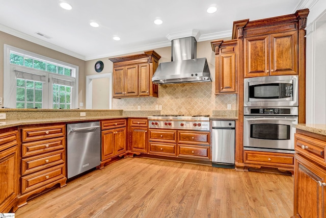 kitchen featuring stainless steel appliances, wall chimney range hood, brown cabinets, light wood-style floors, and crown molding