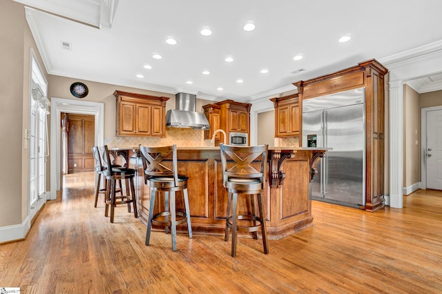 kitchen featuring built in appliances, wall chimney exhaust hood, ornamental molding, tasteful backsplash, and light wood-style flooring