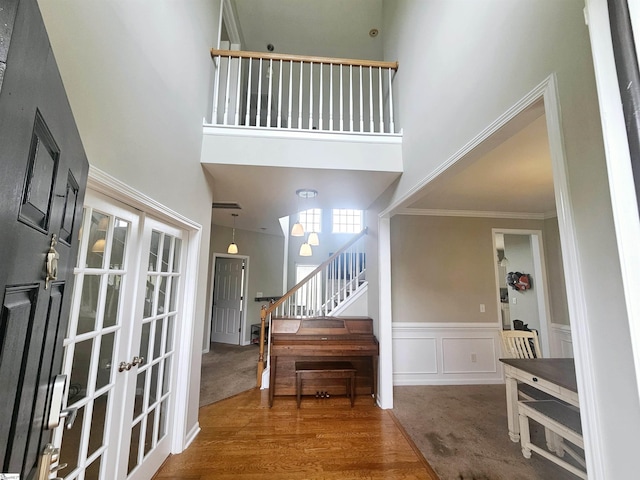 carpeted foyer featuring a towering ceiling, stairway, french doors, a wainscoted wall, and crown molding