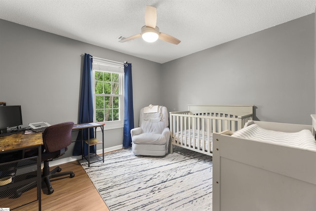 bedroom featuring wood finished floors, an office area, ceiling fan, a textured ceiling, and a crib