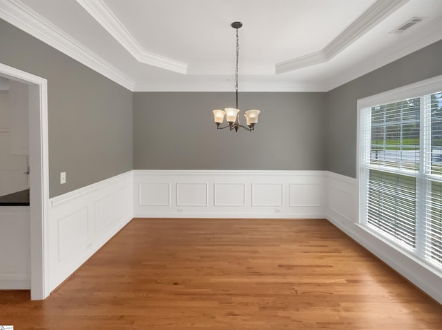 unfurnished dining area featuring a raised ceiling, a chandelier, ornamental molding, light wood-style flooring, and a wainscoted wall
