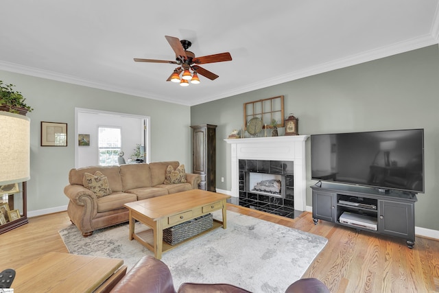 living room featuring a ceiling fan, ornamental molding, light wood-style floors, baseboards, and a tile fireplace