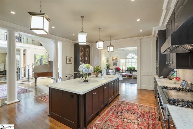 kitchen featuring an island with sink, pendant lighting, ornate columns, wall chimney exhaust hood, and light wood-type flooring