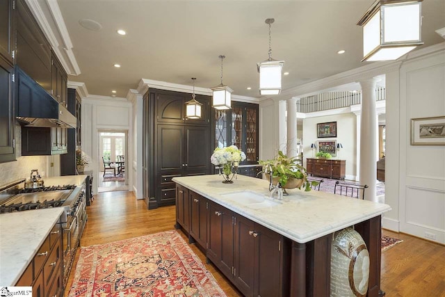 kitchen featuring a center island with sink, light stone counters, crown molding, ornate columns, and pendant lighting