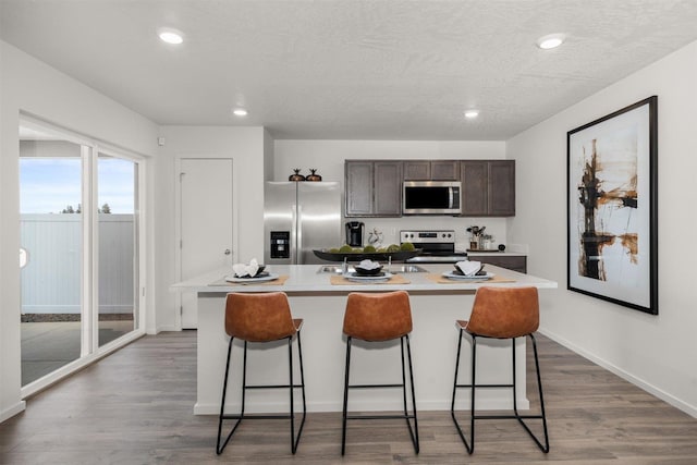 kitchen with a breakfast bar, appliances with stainless steel finishes, a kitchen island with sink, dark brown cabinets, and wood-type flooring