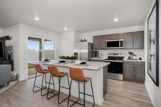 kitchen with light hardwood / wood-style flooring, dark brown cabinets, an island with sink, and appliances with stainless steel finishes