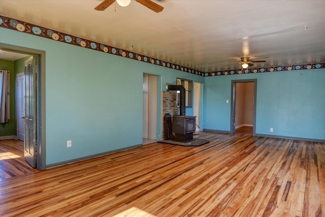 unfurnished living room with a wood stove, ceiling fan, baseboards, and wood finished floors