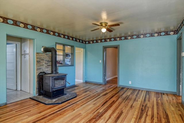 unfurnished living room with a wood stove, light wood-type flooring, baseboards, and a ceiling fan