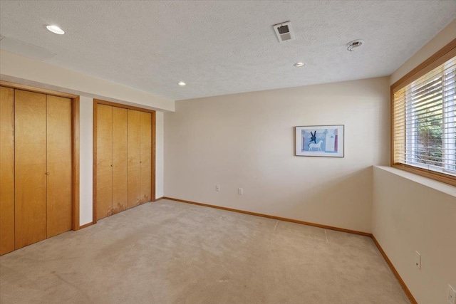 unfurnished bedroom featuring a textured ceiling, light carpet, visible vents, baseboards, and two closets