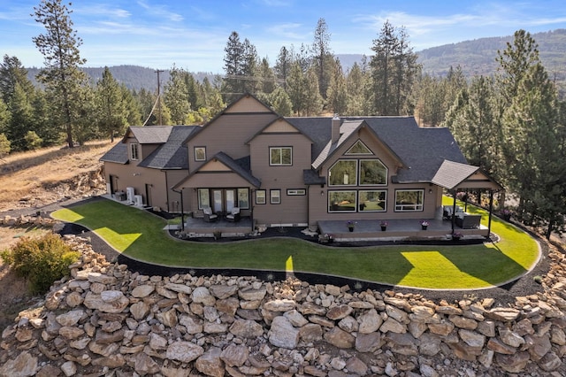 back of property featuring a view of trees, a patio, roof with shingles, a mountain view, and a lawn