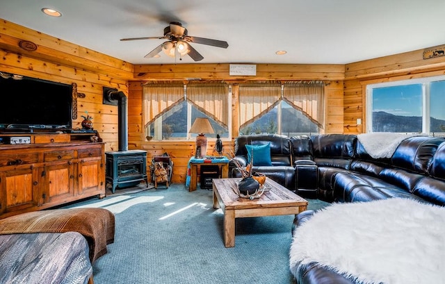 living room featuring wooden walls, ceiling fan, a wood stove, and carpet flooring