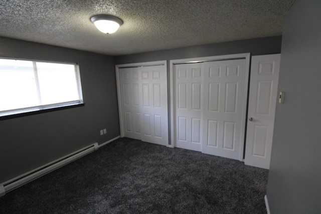 unfurnished bedroom with a textured ceiling, a baseboard heating unit, two closets, and dark colored carpet